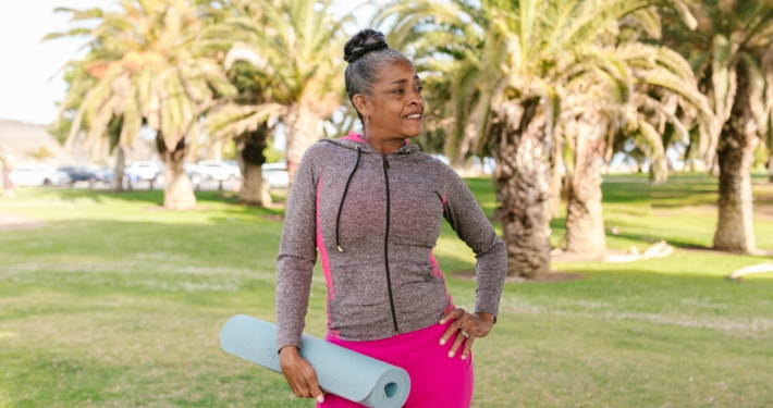 Senior woman smiling with yoga mat in a sunny park, embodying active lifestyle and wellness.